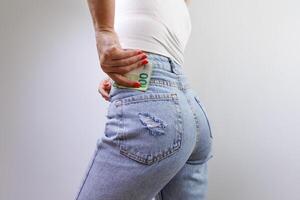 de flicka glider en hundra euro räkningen in i de tillbaka ficka av henne jeans. en flicka i jeans med en euro sedel i henne händer. foto