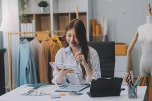 porträtt skön leende asiatisk designer kvinna använda sig av dator i skräddare tyg mode små företag uppkopplad verkstad. ung ägare börja entreprenör. kreativ flicka textil- plagg sme företag begrepp foto