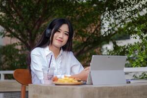 kontor skön asiatisk kvinna i utomhus- Kafé. flicka med en svag leende lyssnande till musik medan skriver håller på med arbete i främre av de bärbar dator ser på de kamera för innehåll eller kampanjer foto