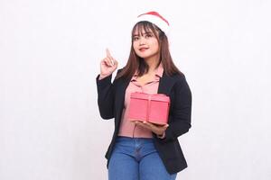 skön asiatisk flicka i glad kostym med santa claus hatt Framställ med jul gåva låda och händer pekande upp på vit bakgrund för befordran, reklam, baner, anslagstavla foto