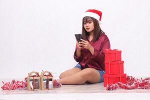 skön ung sydöst asiatisk flicka skriver ett beställa på en grej Nästa till 3 gåva lådor av hindrar på jul bär en santa pannband och röd skjorta på en vit bakgrund för befordran och reklam foto