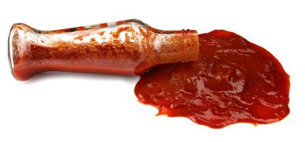 tomat sås . glas flaska med ketchup och en pöl av ketchup spillts runt om den isolerat på en vit bakgrund. foto