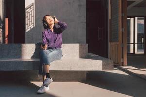 glad ung asiatisk kvinna som lyssnar på musik med hörlurar via smartphone och har kul när hon sitter vid sidan av gatan foto