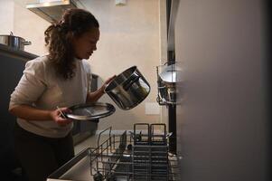 mång etnisk kvinna, hemmafru läser in smutsig maträtt inuti en diskmaskin, håller på med hushållsarbete på Hem kök interiör med minimalistisk design. hushåll. sysslor. hushållning. inhemsk liv och rutin- foto