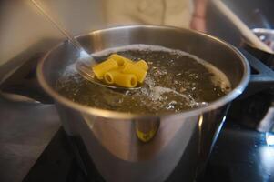 närbild av sätta rå pasta in i kokande vatten i en rostfri stål panorera uppvärmning på ett induktion spis foto