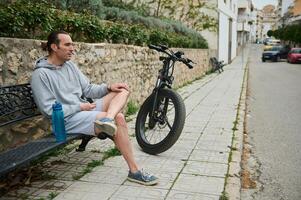ung man cyklist Sammanträde på en bänk nära hans elektrisk cykel, avkopplande efter rida i berg stad foto