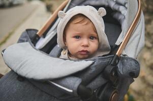 porträtt av ett förtjusande bebis pojke med huva på hans huvud, ser på de kamera, Sammanträde i en bebis sittvagn utomhus foto