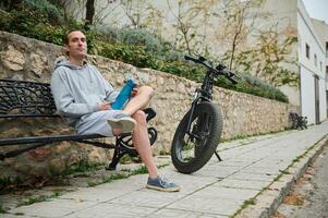 ung caucasian man i grå luvtröja och sporter shorts, Sammanträde på en bänk nära hans hyrd elektrisk cykel, avkopplande i de urban miljö foto