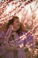 kvinna blomning persika fruktträdgård. mot de bakgrund av en pittoresk persika fruktträdgård, en kvinna i en lång rosa klänning och hatt åtnjuter en fredlig promenad i de parkera, omgiven förbi de skönhet av natur. foto