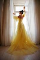 en kvinnas silhuett i en gyllene lyxig klänning mot de bakgrund av en fönster innehar en ridå med henne händer. elegant lady i en gul lång silke klänning med bar tillbaka, tillbaka se. foto