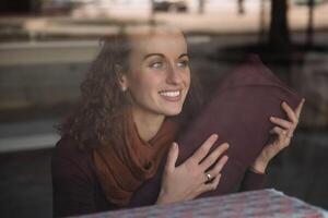 lockigt hår kvinna sett genom en glas skyltfönster, innehav en bit av vinröd tyg och leende varmt foto