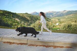 full längd sida porträtt av en söt barn flicka löpning medan gående henne hund på koppel på de natur utomhus foto