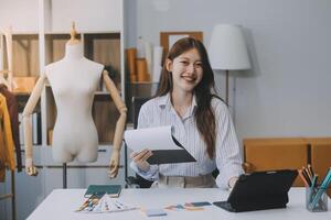 porträtt skön leende asiatisk designer kvinna använda sig av dator i skräddare tyg mode små företag uppkopplad verkstad. ung ägare börja entreprenör. kreativ flicka textil- plagg sme företag begrepp foto