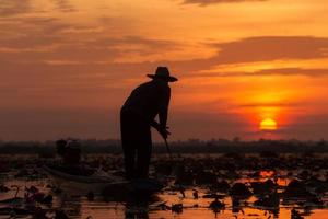 siluettfiskaren som bär hatt och arbetar på en båt under soluppgången vid sjön Udon Thani, Thailand.