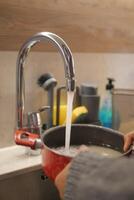 en person är använder sig av de kök handfat till tvätta en pott med vatten foto