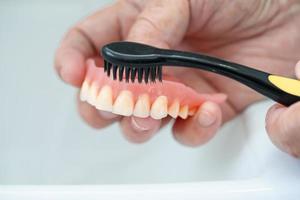 asiatisk senior eller äldre gammal kvinna patienten använder tandborste för att rengöra delvis protes av ersättningständer.