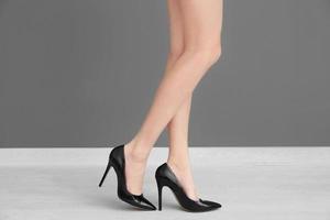ben på ung kvinna i skor med höga klackar som står nära grå vägg