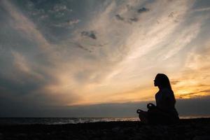 siluett av en kvinna i meditation poserar på havsstranden under surrealistisk solnedgång på havsbakgrund och dramatisk himmel foto