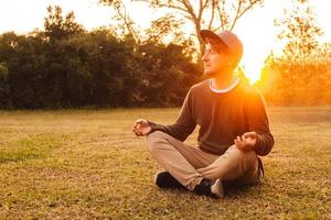 man i en meditativ position sitter på en gräsmatta på en bakgrund av skog och solnedgång foto