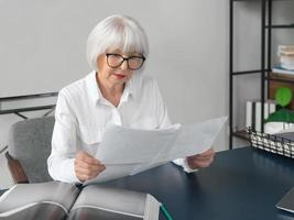 trött senior vacker grått hår kvinna i vit blus läser dokument på kontoret. arbete, seniorer, frågor, hitta en lösning, uppleva koncept foto