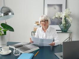 trött senior vacker grått hår kvinna i vit blus läser dokument på kontoret. arbete, seniorer, frågor, hitta en lösning, uppleva koncept