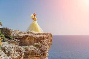 kvinna gul klänning hav. sida se ung skön sensuell kvinna i gul lång klänning Framställ på en sten hög ovan de hav på solnedgång. flicka i natur mot de blå himmel foto