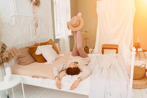 livsstil begrepp. en ung medelålders kvinna i en Tröja lögner på de säng på henne tillbaka, skrattar och innehar en brun hatt på henne fötter. foto