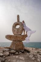 en kvinna står på en sten skulptur tillverkad av stor stenar. hon är klädd i en vit lång klänning, mot de bakgrund av de hav och himmel. de klänning utvecklas i de vind. foto