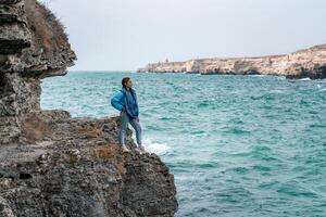 en kvinna i en blå jacka står på en sten ovan en klippa ovan de hav och utseende på de rasande hav. flicka resande vilar, tänker, drömmar, åtnjuter natur. fred och lugna landskap, blåsigt väder. foto