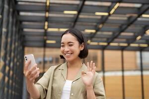 Lycklig asiatisk flicka vågor på henne smartphone kamera, säger Hej till vän på chatt, kallelse någon, stående på gata foto