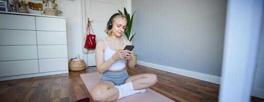 porträtt av ung blond kvinna i hörlurar, vändning på yoga, träna app på smartphone, välja musik på mobil telefon Ansökan, Sammanträde på sudd matta i rum foto