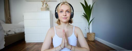 bild av ung kvinna lyssnar till yoga podcast för nybörjare, innehar händer i namaste tecken, bär hörlurar, mediterar foto