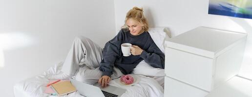 porträtt av ung kvinna, studerande studerar i henne säng, avkopplande medan framställning läxa, äter munk, använder sig av bärbar dator i sovrum och dricka te foto