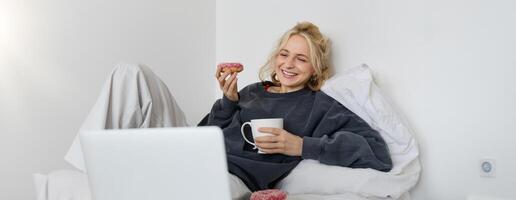 livsstil och människor begrepp. Lycklig blond kvinna liggande i säng med mat, dricka te och äter munk, vilar på Hem, utgifterna tid ensam, tittar på TV visa foto