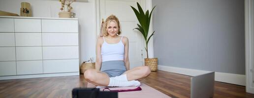 porträtt av ung kondition instruktör, vlogger som visar övningar på kamera, inspelning själv, Sammanträde på matta med bärbar dator, håller på med träna, förklara yoga rörelser till följare foto