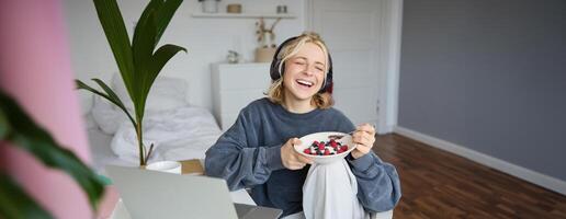 porträtt av Lycklig ung blond kvinna, Sammanträde i en rum, tittar på film på bärbar dator och äter friska frukost, dricka te, vilar på helgen foto