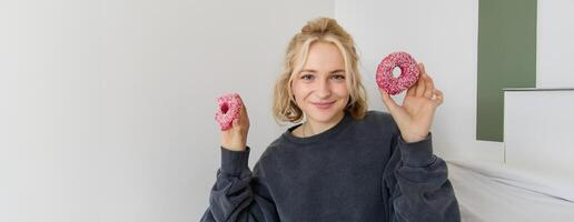 stänga upp porträtt av Lycklig, söt blond kvinna, innehav munk, äter ljuv, utsökt bekvämlighet mat, som visar efterrätt på kamera foto