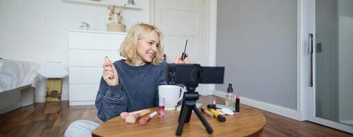 porträtt av ung leende kvinna i henne rum, inspelning på kamera, livsstil vlog för social media, innehav mascara, granskning henne smink skönhet Produkter, som visar på vilket sätt till använda sig av kosmetika foto