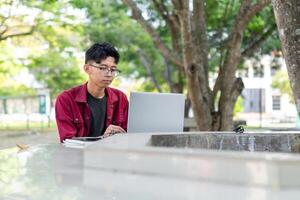 porträtt av asiatisk högskola studerande använder sig av bärbar dator ser till kamera. en man arbetssätt med en bärbar dator dator på campus foto