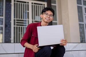 porträtt av asiatisk högskola studerande använder sig av bärbar dator ser till kamera. en man arbetssätt med en bärbar dator dator på campus foto