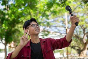 asiatisk man inspelning och talande till kamera. ung man vlogging och tar selfie med verkan kamera på offentlig parkera. foto