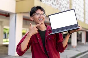 ung asiatisk man bär röd skjorta leende och som visar vit bärbar dator skärm för annons. begrepp av människor livsstil. Lycklig man innehav tom bärbar dator skärm med suddig bakgrund. foto