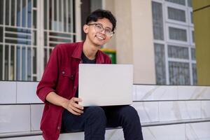 porträtt av asiatisk högskola studerande använder sig av bärbar dator ser till kamera. en man arbetssätt med en bärbar dator dator på campus foto