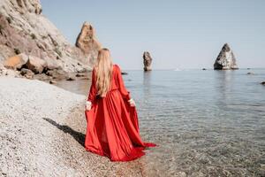 kvinna resa hav. Lycklig turist i röd klänning njut av tar bild utomhus för minnen. kvinna resande Framställ på de sten på hav bukt omgiven förbi vulkanisk berg, delning resa äventyr resa foto