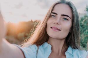 bärnsten gyllene timme stänga upp av en kvinna badade i gyllene timme ljus framställning selfie på solnedgång i sommar parkera. Lycklig turist njut av tar selfie Foto utomhus för minnen