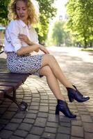 skön mitten ålder kvinna i 70-talet, 80s stil kläder på en bänk i en solbelyst aveny foto