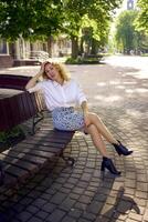 skön mitten ålder kvinna i 70-talet, 80s stil kläder på en bänk i en solbelyst aveny foto