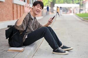 en glad asiatisk manlig högskola studerande sitter på de gata förbi de campus byggnad, vinka hans hand. foto