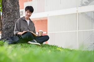 en Lycklig, avslappnad asiatisk manlig högskola studerande åtnjuter läsning en bok under de träd i de campus parkera. foto