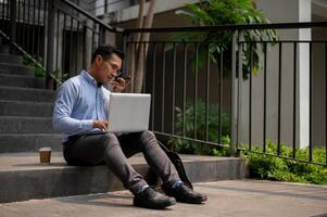 en affärsman sitter på steg, sändning röst meddelande via hans smartphone medan arbetssätt på hans bärbar dator. foto
